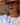 A close-up lifestyle shot of a woman wearing a gold chain necklace with a blue bear charm and a small gold padlock pendant, posing in front of a brick wall.